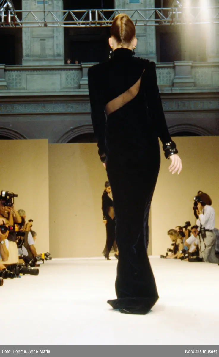 Modevisning. Modell i svart långklänning, armband och håret i svinrygg, fotograferad med ryggen mot kameran. Från Balmain.