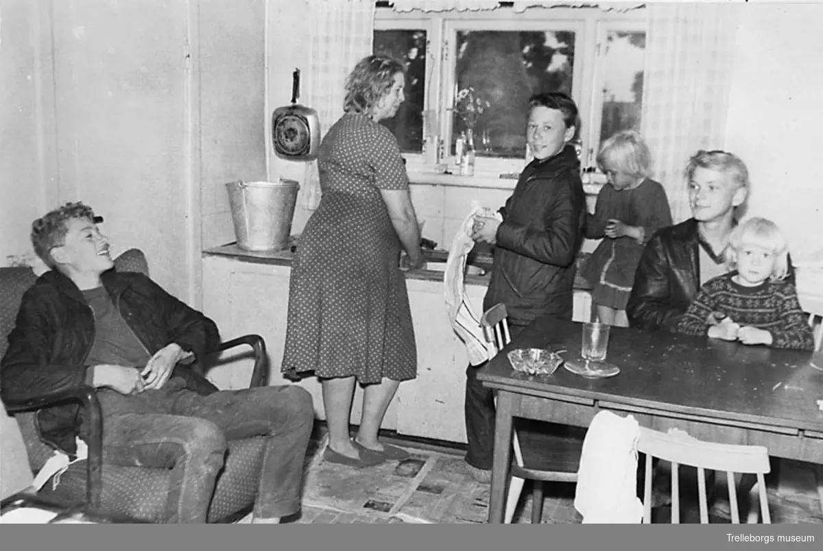 En familj i Fuglie år 1965. Mor diskar och sonen torkar disken. På golvet ligger det tidningar.