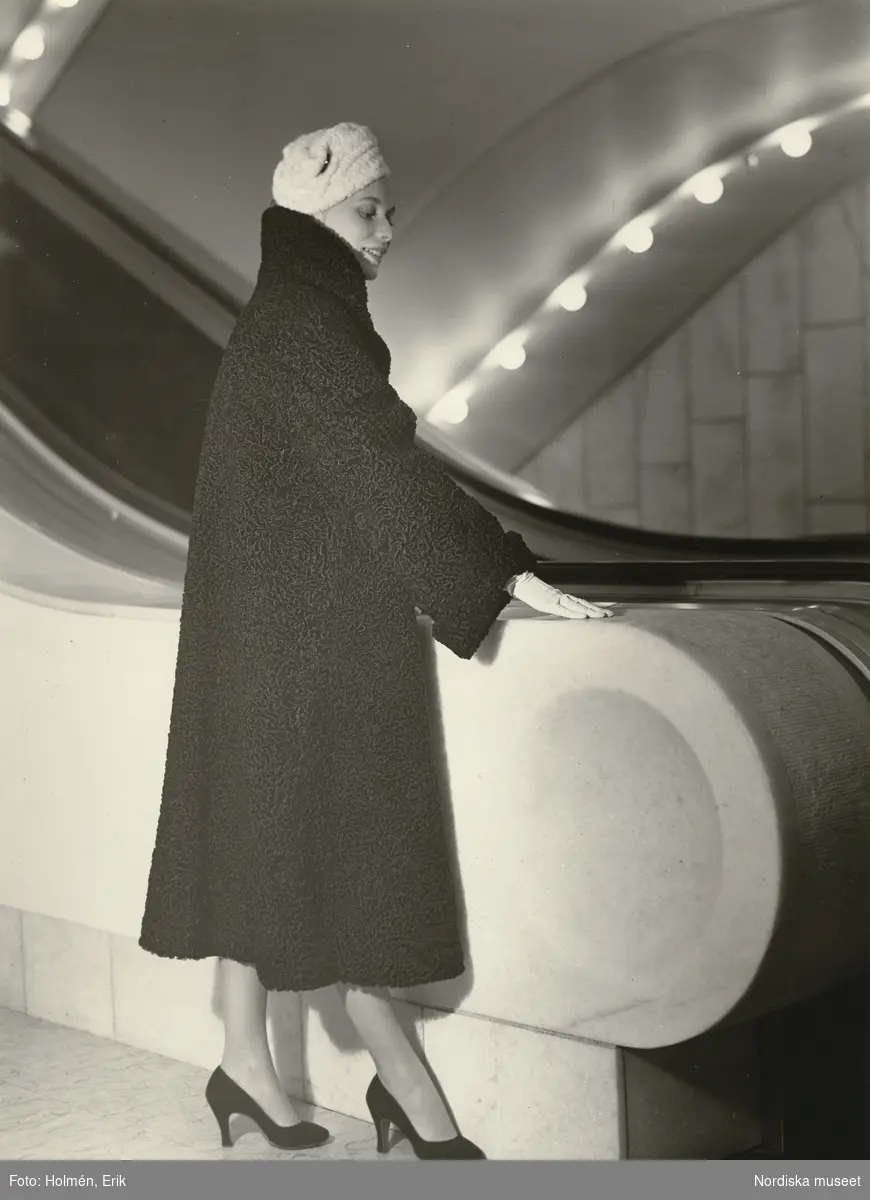 Nordiska Kompaniet. Kvinna i persianpäls vid en av NK:s rulltrappor. Ljus hatt, ljusa handskar. Text på baksidan: "Pälsar 1940-talet."
