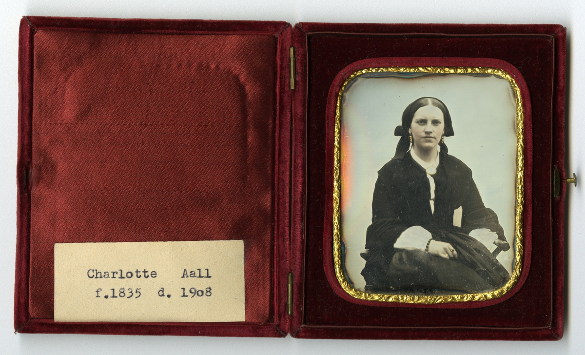 Unika. Daguerrotypi i etui. Etui dekket med rød fløyel. To små hengsler og knapp til å åpne. Innvendig portrett av sittende kvinne, gullkant med reileffdekor rundt fotogarfiet, avrundede hjørner. Kvinnen er Charlotte Aall (1835-1908).