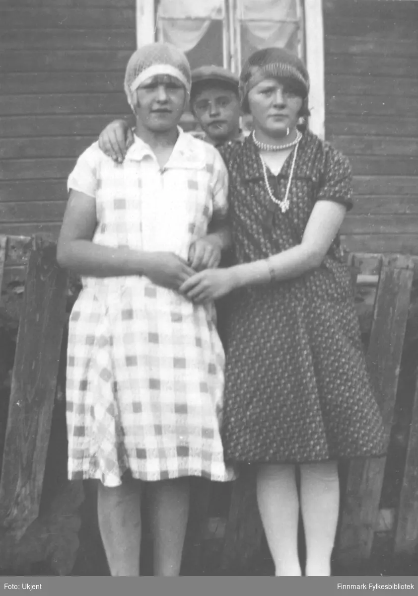 Gruppeportrett at to jenter som poserer i kjoler og tidsriktige hodeplagg fra 20-30.årene. En litt yngre gutt titter over skulderen på jentene. Jenta til høyre har perlekjede om halsen. Navn ukjent. Muligens tatt i Vadsø.