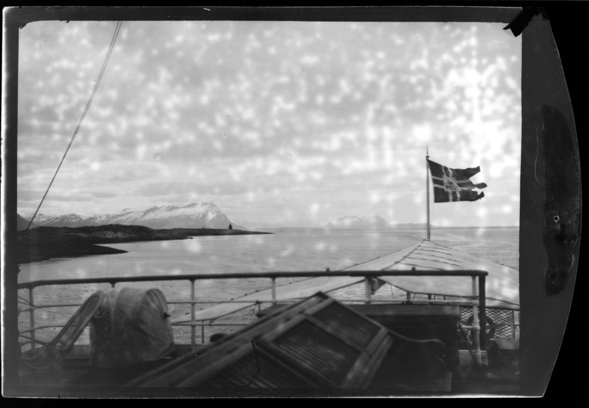 Bildet er tatt fra hurtigruten mot fastland. Ombord på akterdekk står en dekkstol. Norsk flagga er heist opp. På fastland (langt borte) står en fyr.