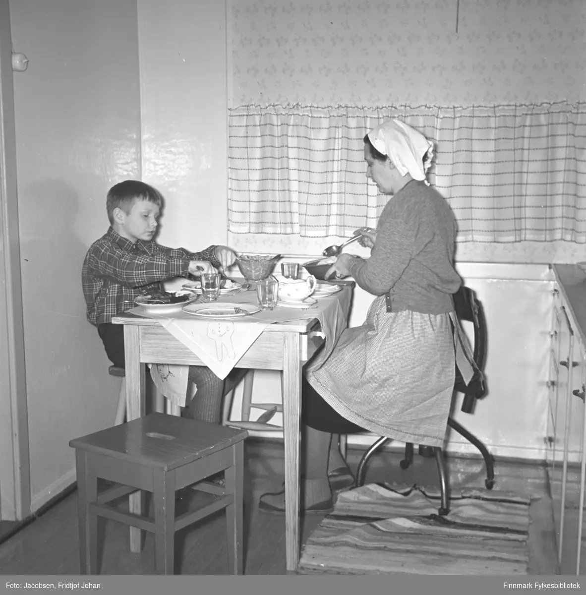 Aase og sønnen Arne Jacobsen ved middagsbordet på kjøkkenet i Storvannsveien. Arne har en rutet skjorte og ganske lys bukse på seg. Aase har et mørk skjørt, med et lyst forkle over og en litt mørkere jakke. På hodet har hun et hvitt skaut/tørkle. Arne sitter på en trekrakk og Aase på en kontorstol. Tallerkener, glass, skål og bestikk ligger på bordet mellom dem. En krakk og en rye ses på gulvet og en liten del av kjøkkeninnredningen ses helt til høyre på bildet. En lys gardin henger foran vinduet.