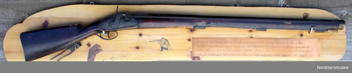 Rifle som er montert på plakett med medfølgende kuletang. På plaketten står opplysninger inngravert på plate og med tegning av bjørnejakt.