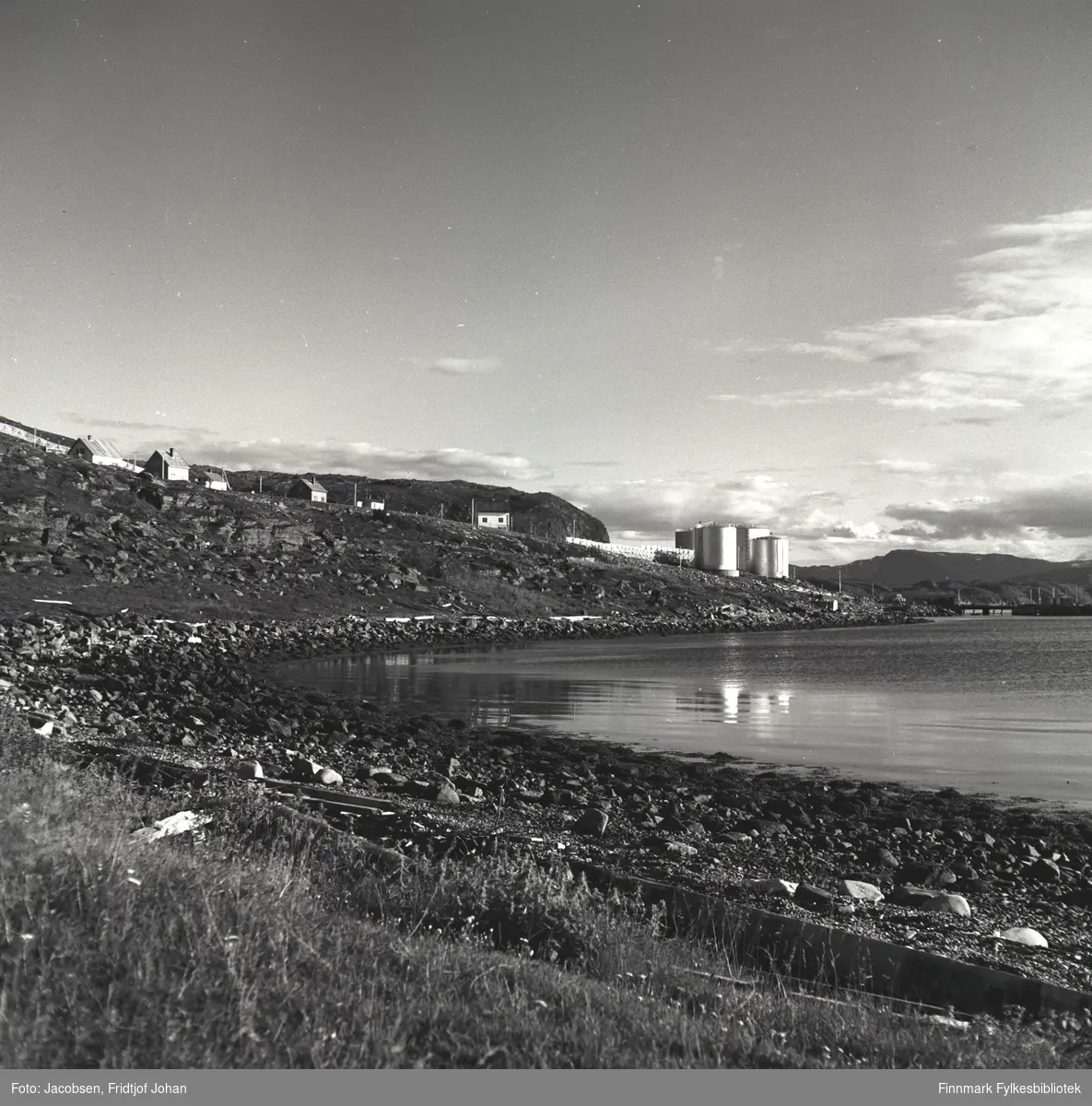 Shells tankanlegg på Stornes i Rypefjord. Noen bygninger står langs Fjordaveien som går forbi Storneset. Der står også endel el-stolper på rekke og rad. En snøskjerm ses helt til høyre på bildet. Noe gress står i terrenget rett foran fjæra og sjøen er ganske stille i havna.