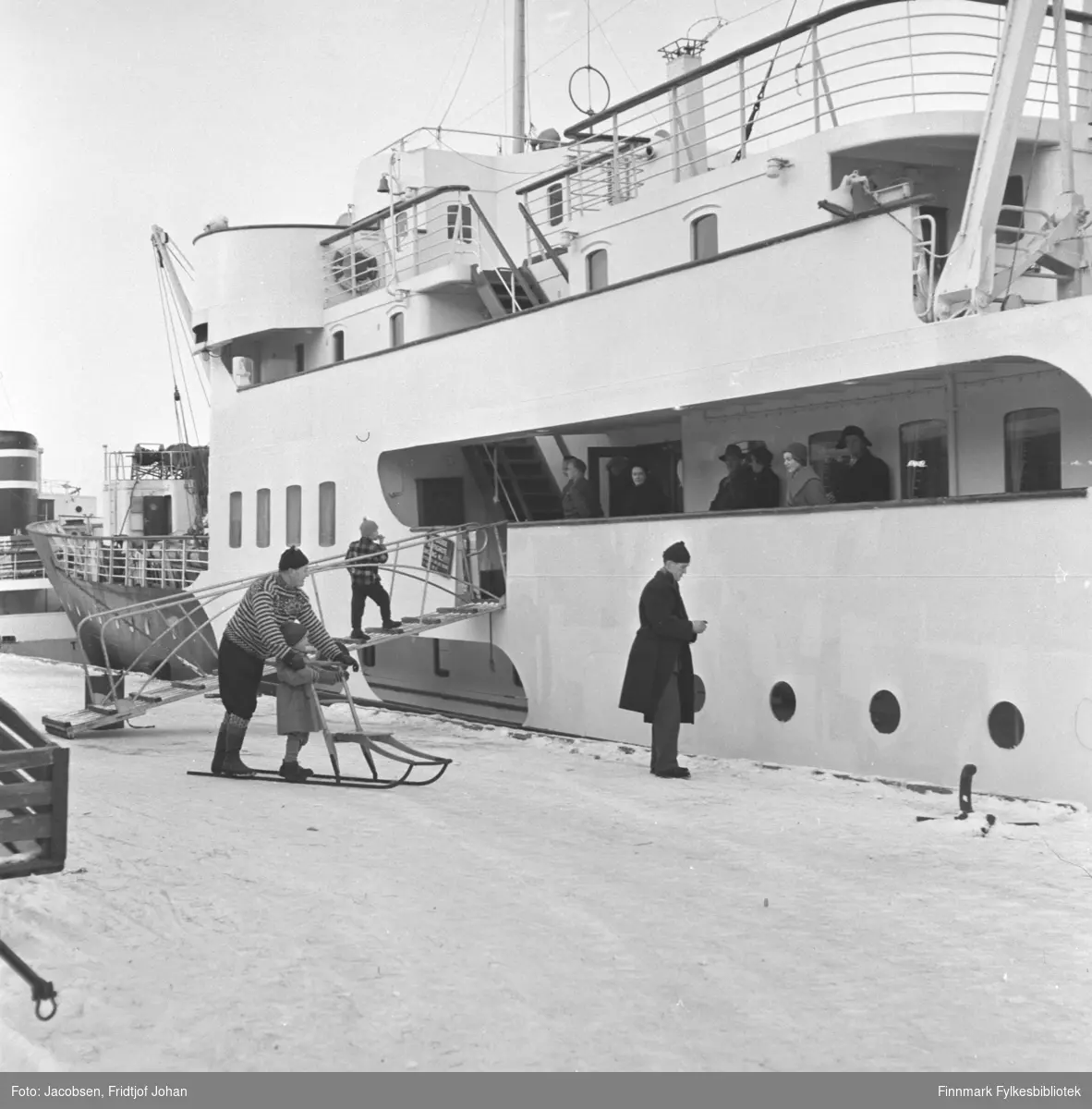 Hurtigruten Ragnvald Jarl klar for avgang fra Hammerfest havn. Flere personer står på dekk og en gutt er på tur opp landgangen. En mørkkledt person står på kaikanten og en mann med spark er like bak. En jente står på meiene til sparken. En liten del av et overbygg og en skorstein til en annen båt ses til venstre på bildet.