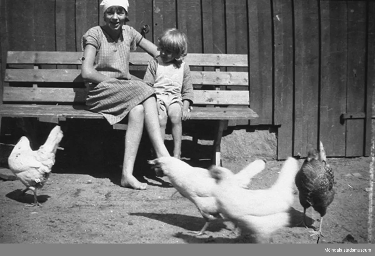 Sommar i Lindome i början av 1940-talet. En kvinna och ett barn sitter på en bänk. Framför dem går några hönor.