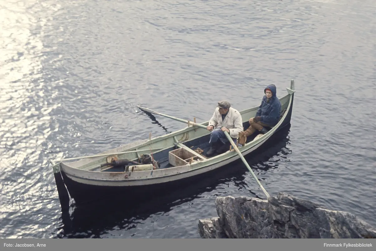 Arne Nakken og Aase Jacobsen på tur i Nordlandsbåt. Han med årene og iført lys jakke, gummistøvler og lue. Aase med mørk jakke, hetten på og hansker. I båten ligger en fiskekasse. Det er stille på sjøen og solskinn.