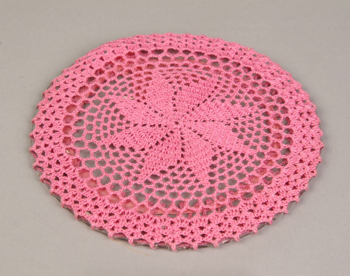 Rund glassplate med en heklet rosa brikke tredd over. Den er festet med en rosa sløyfe på den ene siden.