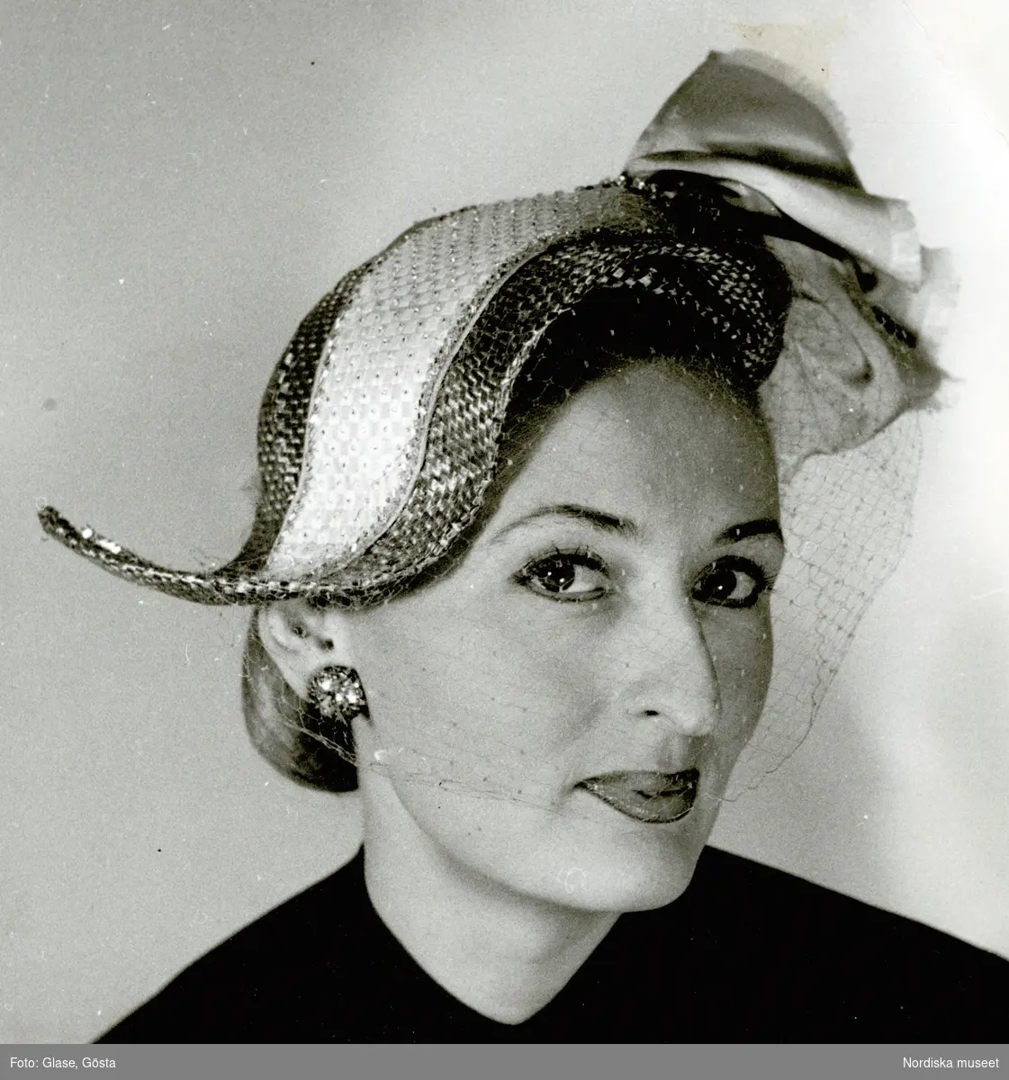 Kvinna i hatt med garnering och flor, samt örhänge och håret uppsatt i knut i nacken.