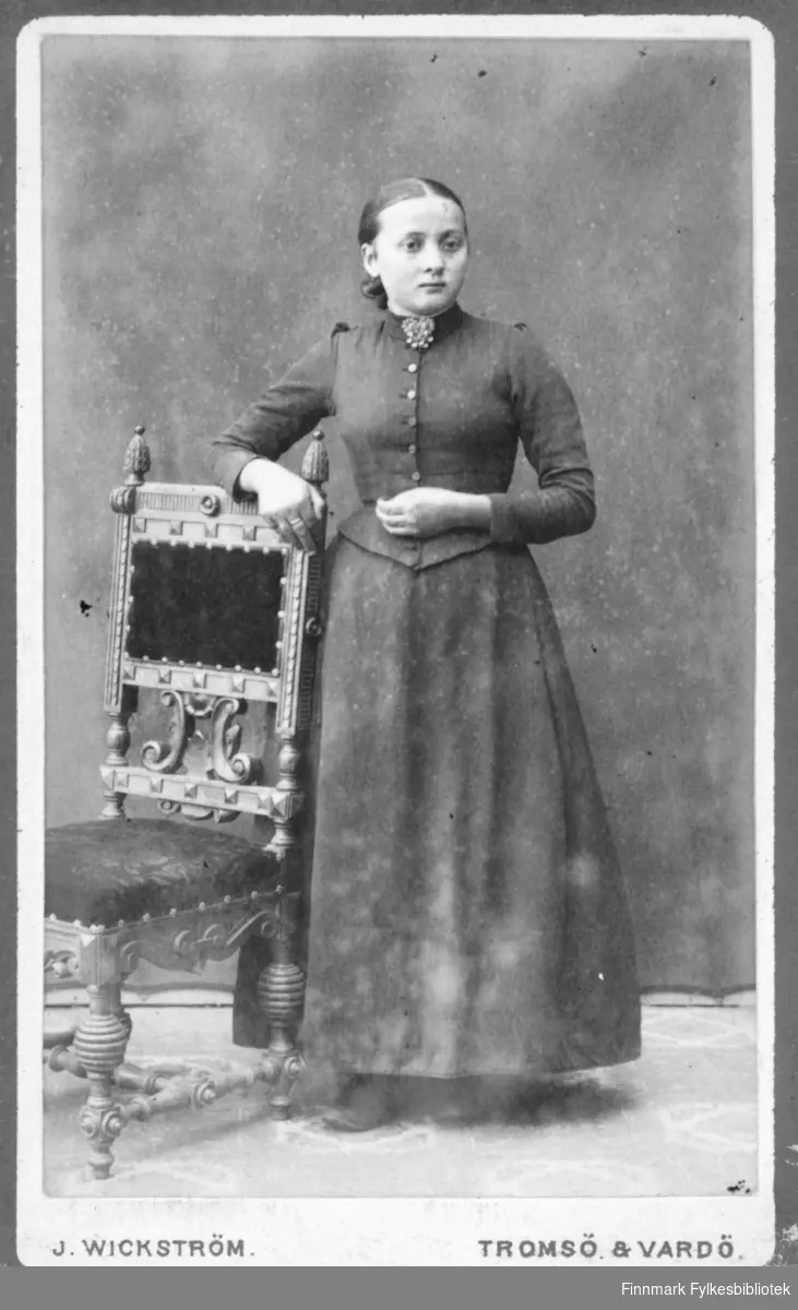 Portrett av ei ung jente, kanskje konfirmasjonsbilde, hun står med en hånd over ryggen på en stol iført kjole og brosje i halsen. Hun har en stor ring på høyre langfinger. Albumet med bildet kommer fra Ekkerøy, kanskje hun bodde der.