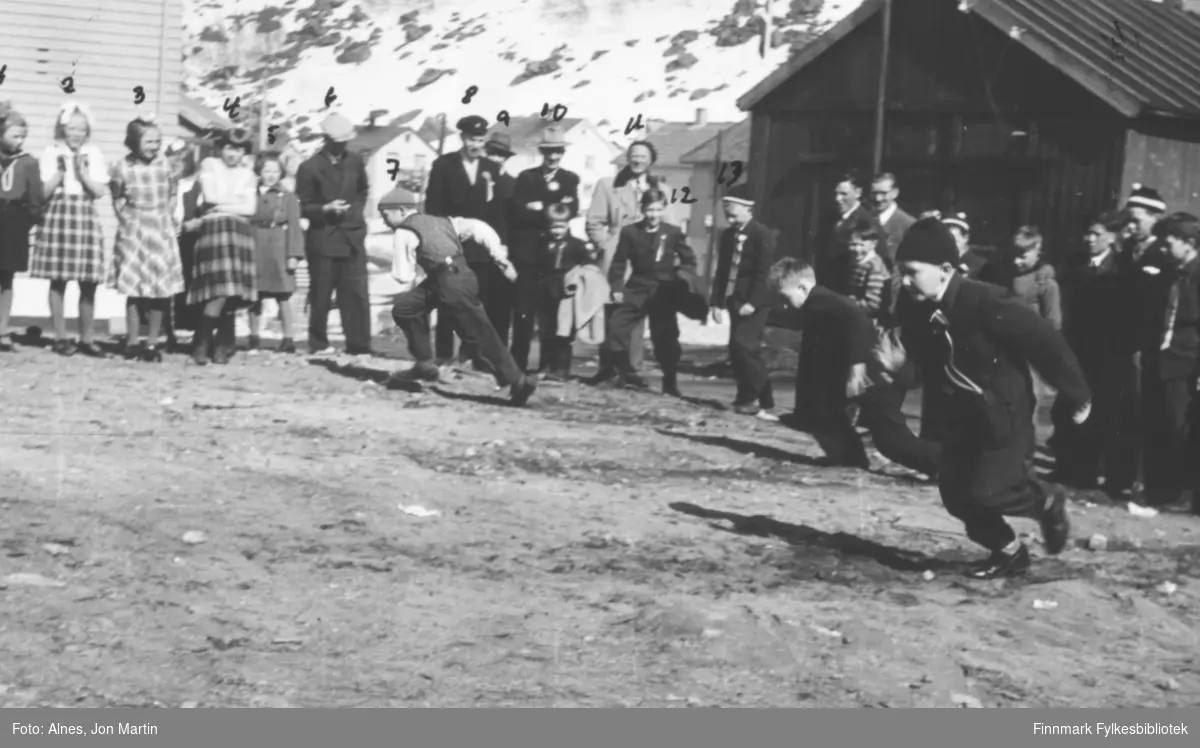 Barneleker 17. mai 1954 i Øksfjord. Tre-fire gutter er midt i leken, omgitt av ivrige tilskuere. Bygningen til venstre var messa, til høyre lekebrakka