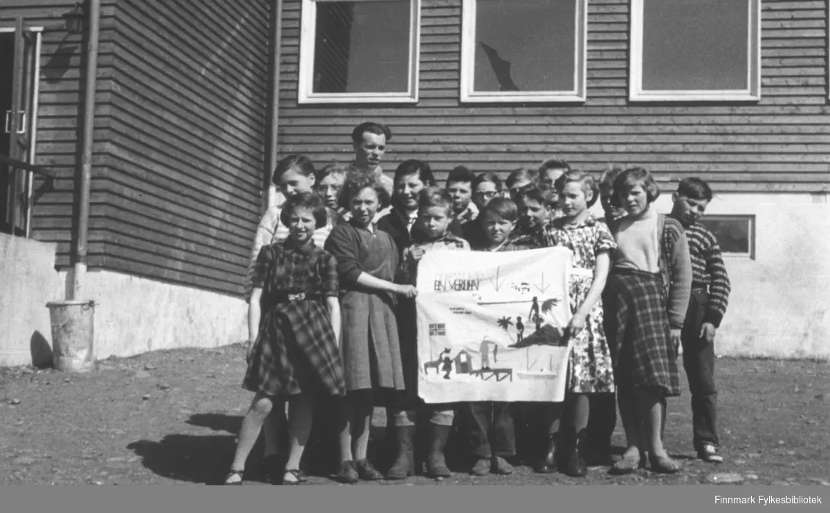 4. klasse ved Øksfjord barneskole fotografert foran skolen sammen med læreren. De holder fram et fint tekstilbilde de har laget, med teksten 'En verden'