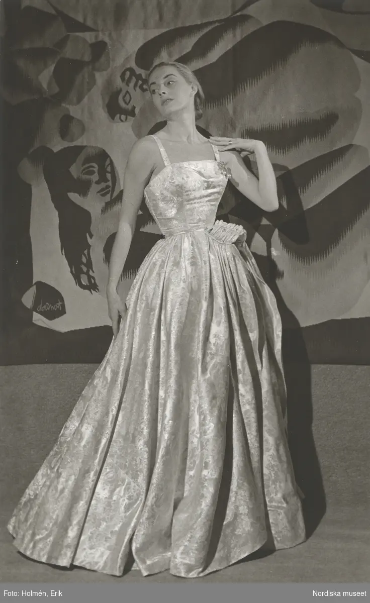 Modell i aftonklänning, framför textilkonstverk. Modell efter Madame Grès.
