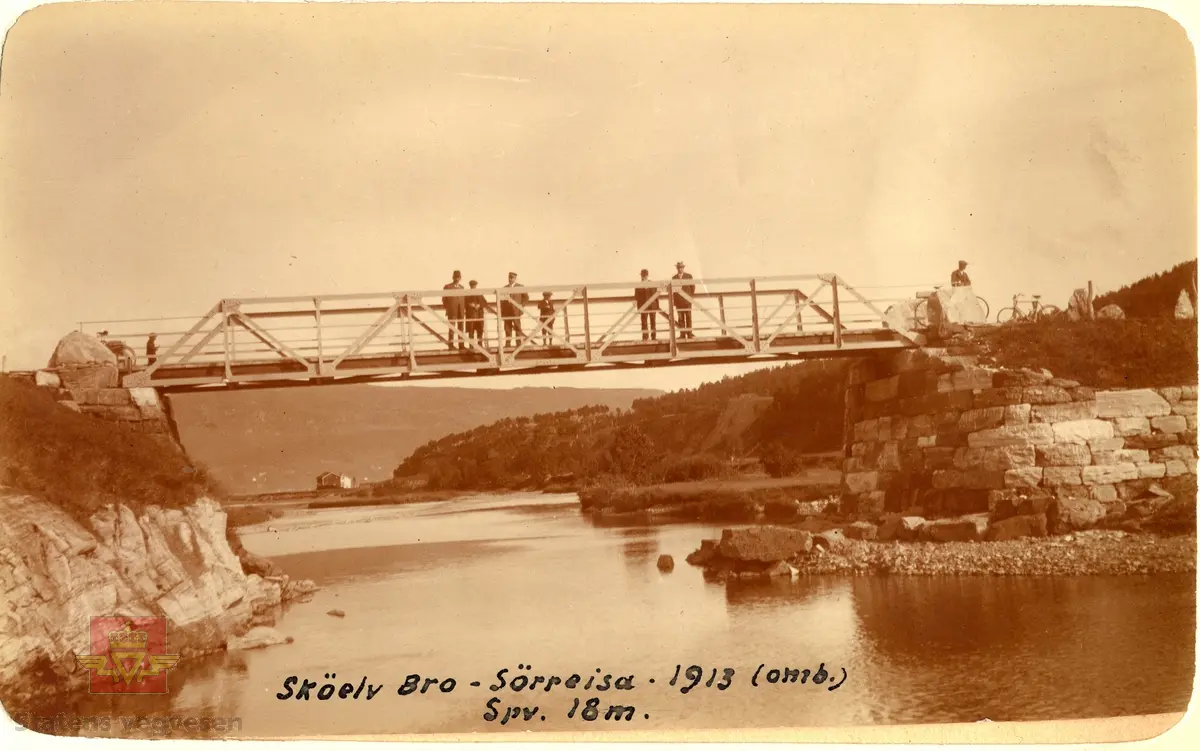 Skøelv bru i Sørreisa. Ombygd i 1913. Spennvidde 18 meter. En gruppe menn og to barn med sykler står på brua. Stabbesteinsrekkverk på bruendene.