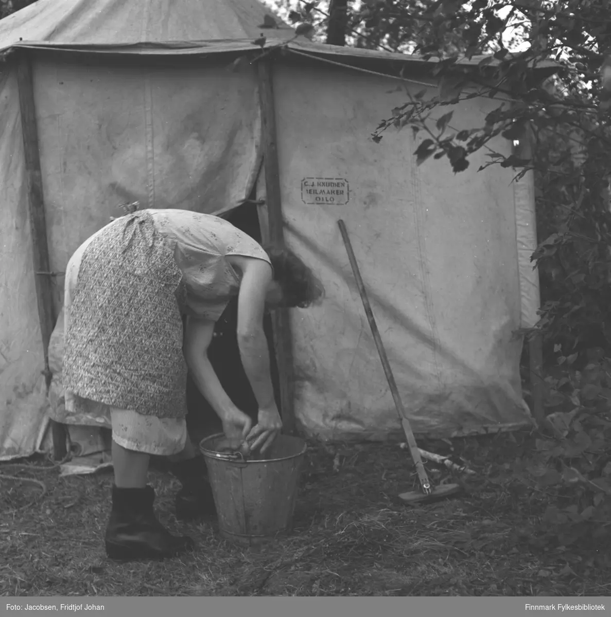 En dame står bøyd over en vaskebøtte utenfor et telt, det kan være Aase Jacobsen. Stedet er ukjent.