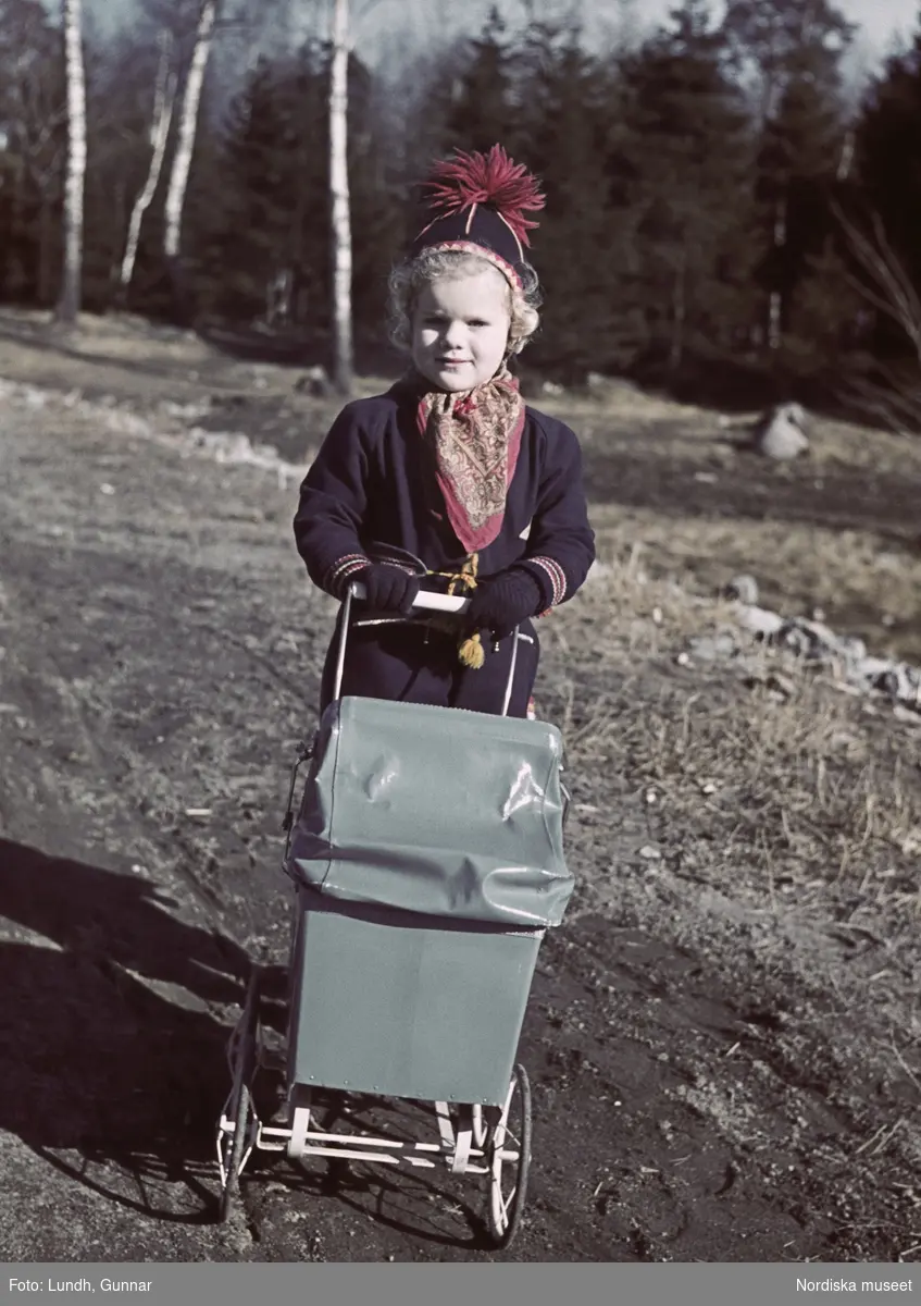 Barn i samisk-inspirerad jacka med bälte, tofsmössa och snusnäsduk i halsen, går med dockvagn ute i naturen.