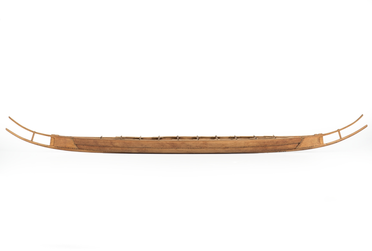 Modell av oxelträ, rekonstruktion av Hjortspringsbåten, utgrävd 1921, daterad till 200-300 f.Kr. Klamparna på borden utsparade i samma stycke. Spanten av piasava. Sammansyningen av borden, kölplankan, stävarna och spanten gjord med svartoxiderad suturtråd. Träet svagt betsat.
Lösa detaljer: 20 st paddlar, 1 st.styråra, 1 st. öskar.