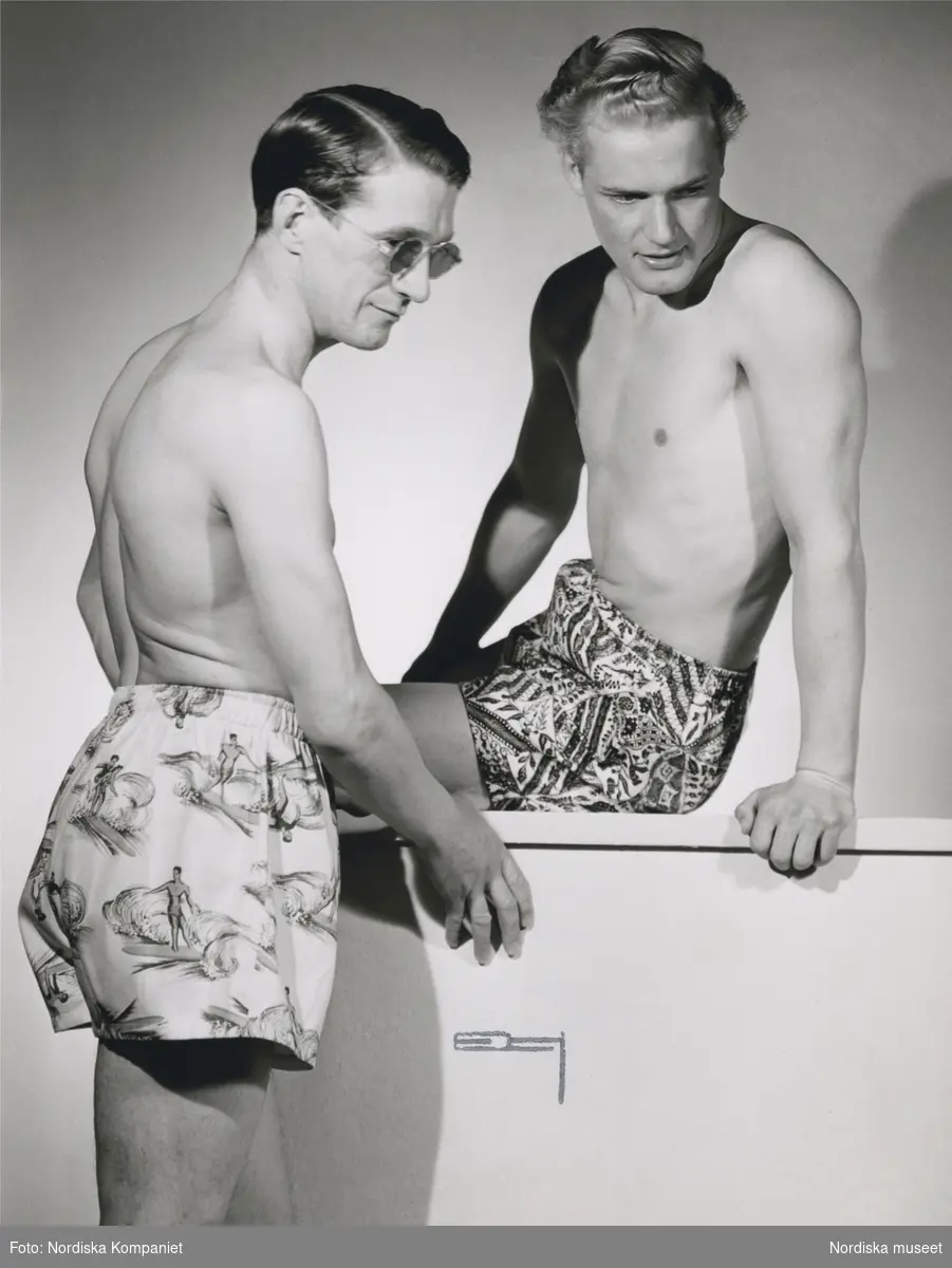 Två män i bar överkropp och badbyxor. Den ena badbyxan med mönster av surfande figurer och höga vågor. Nordiska Kompaniet. Från Nordiska Kompaniets kampanj med Californiakläder i maj 1947.