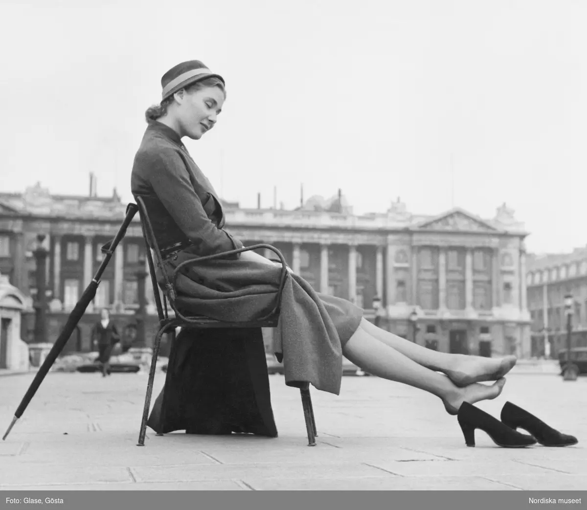 Kvinna i klänning och hatt sitter på en stol vid ett torg och har tagit av sig sina pumps. Mot stolsryggen står ett paraply lutat.