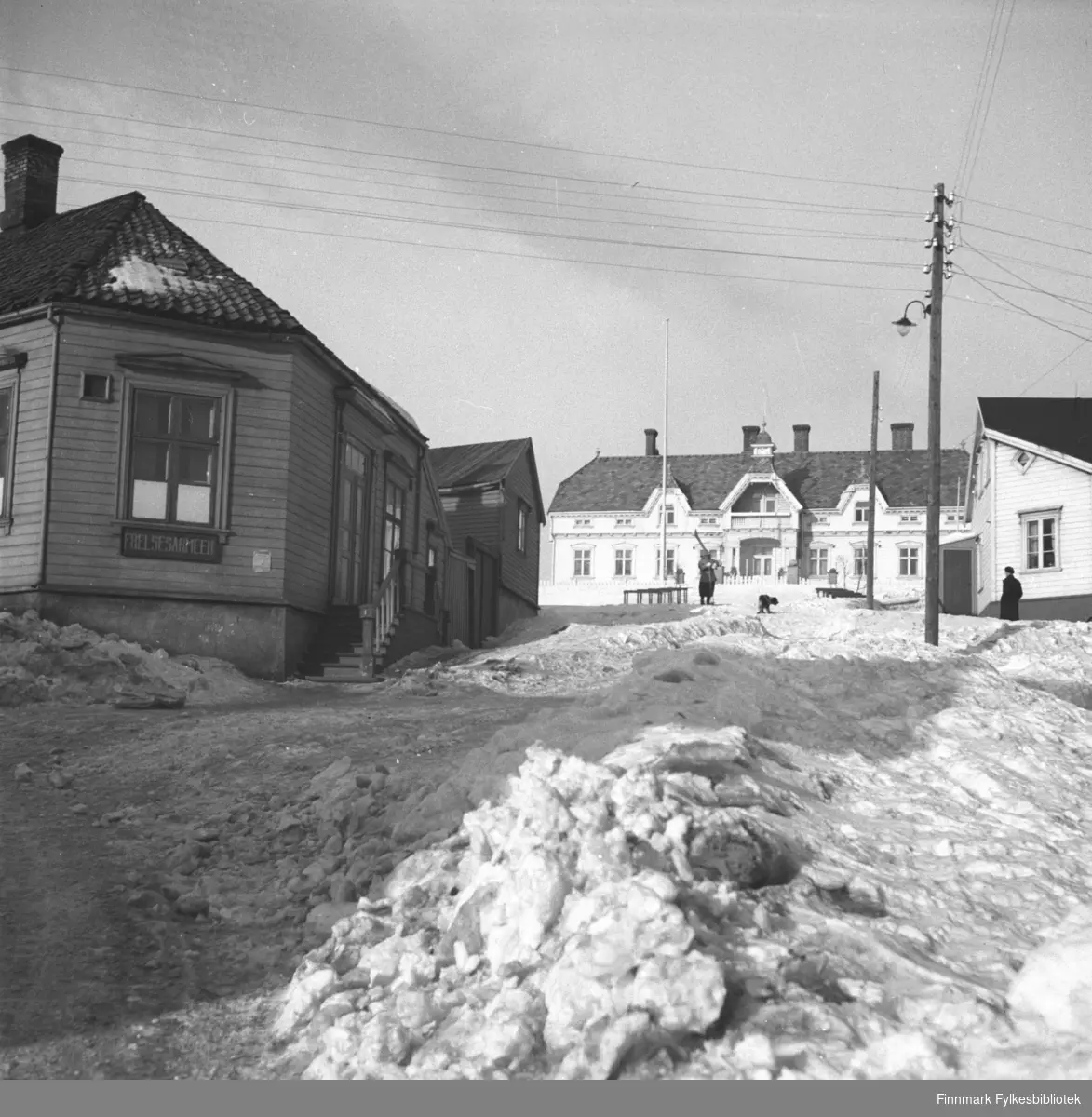 Foto fra Vadsø, avnTollbodgaten, også kalt Apotekerbakken. Til venstre sees Frelsesarmeens lokale, i enden sees Fylkesgården, også kalt Amtmannsgården. På høyre side i bildet står Moes hus. Bildet er muligens tatt i 1934. Fotograf ukjent.