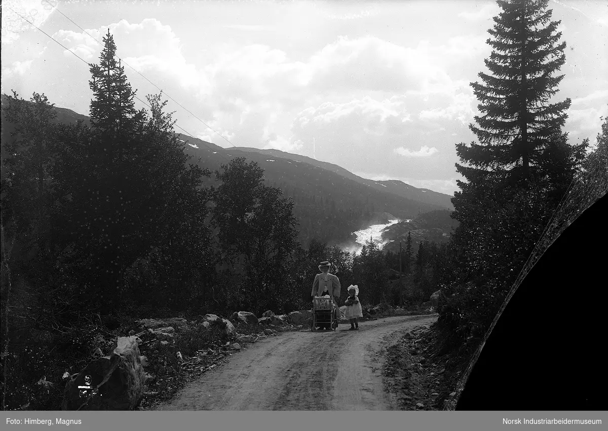 Kvinne og jente gående på vei kledd i pentøy, kvinnen triller på en vogn, muligens en barnevogn. Elv og fjell i bakgrunnen.