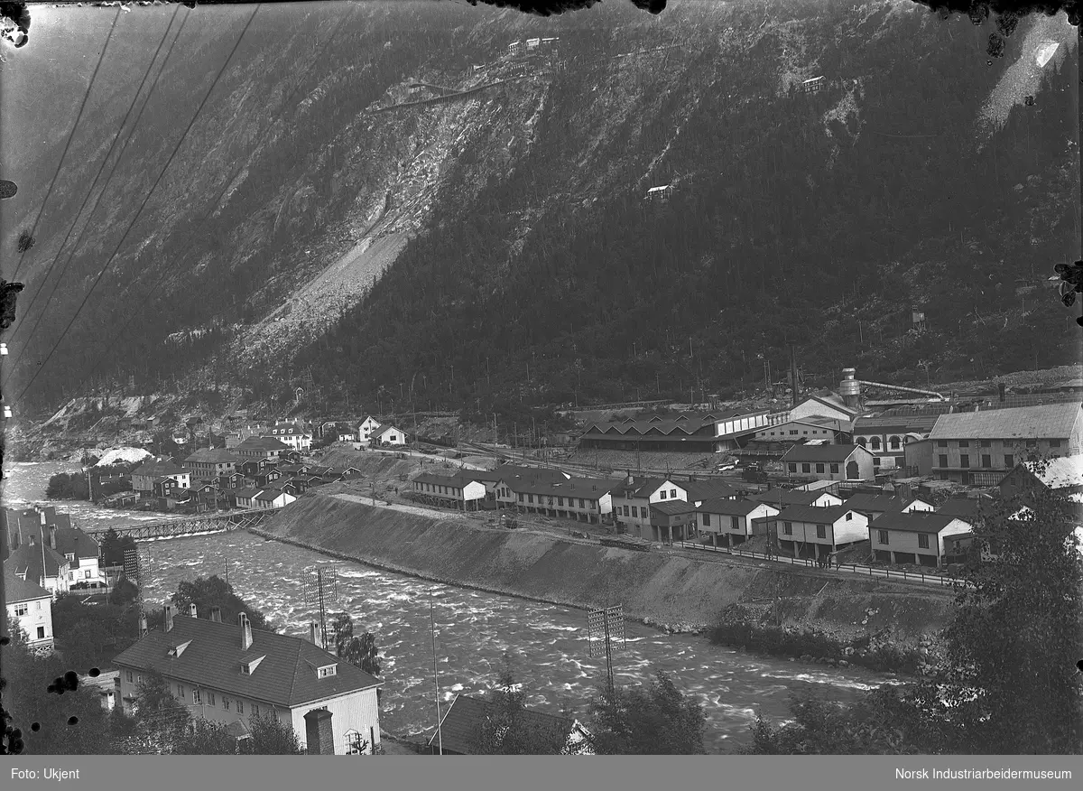 Brakkebyen nord og vest for Rjukan stasjon. Pakkhus på stasjonstomten. Industribygninger på sørsiden av brakkebyen. Boliger i fjellsiden mot sør. Rjukan varelager og boligbebyggelse i Bråvold.