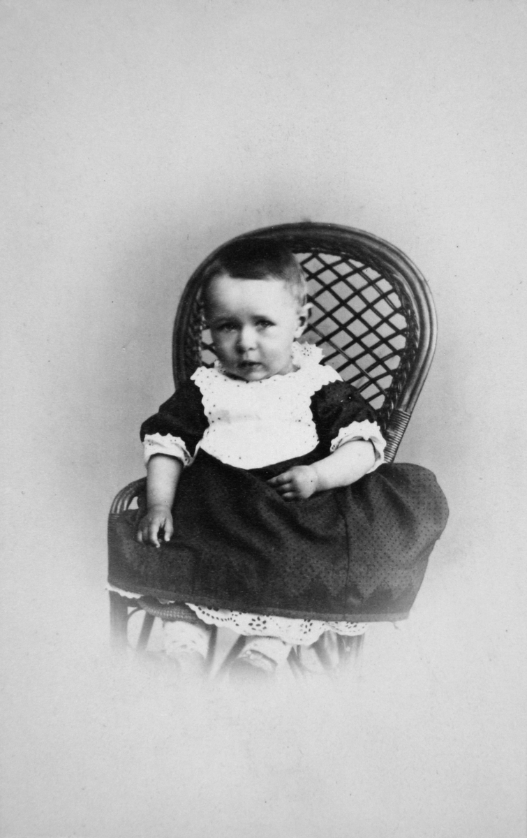 Christian Thams 1 år. Sitter på en barnestol iført mørk kjole med  hvitt skjortebryst.