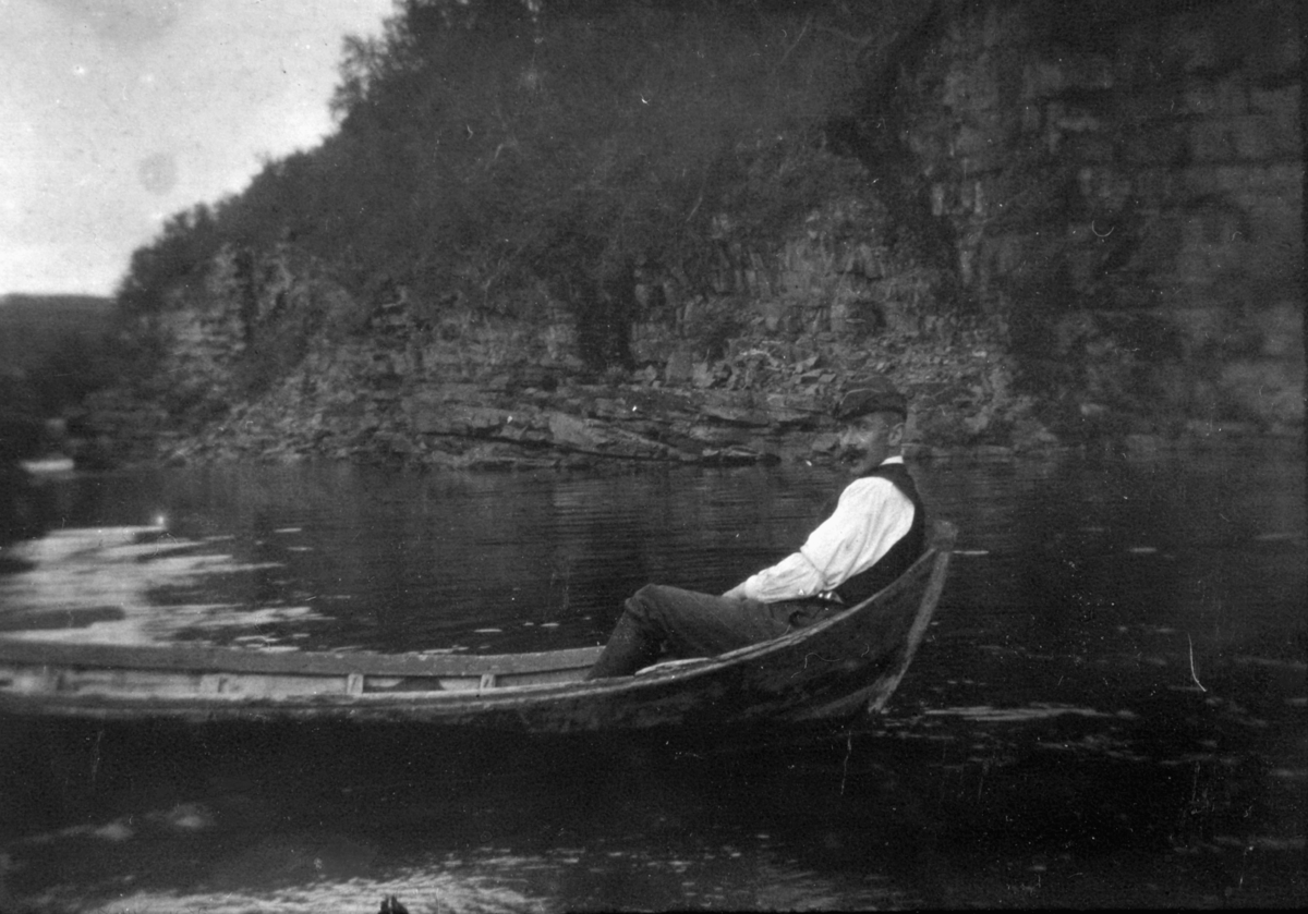 En mann sitter i en båt. Det er muligens Lauritz Lassen Brodtkorb. Båten kan være en elvebåt. Han har på seg skjorte, vest, hatt, bukser og støvler. Lauritz har bart. I bakgrunnen kan man se fjellvegg. Båten ligger rolig på vannet.