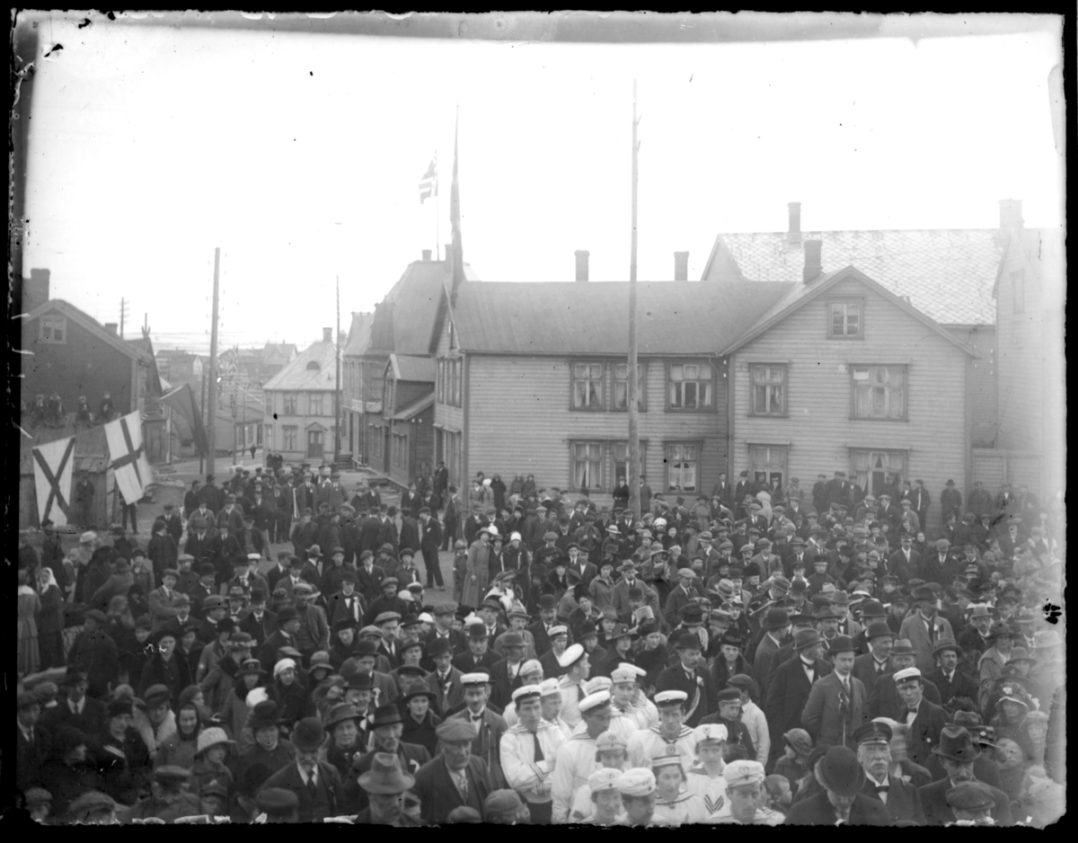 Antakelig 17. maifeiring i Vardø det flagges og en stor folkemengde er samlet. Midt i bildet en gruppe kledd i hvite uniformer, det ser ut til å være et sangkor