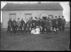 Gruppebilde tatt utenfor en lav bygning med torvtak. Menn, k