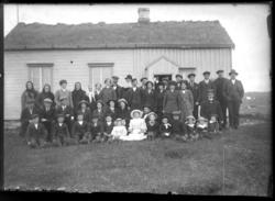 Gruppebilde tatt utenfor en lav bygning med torvtak. Menn, k