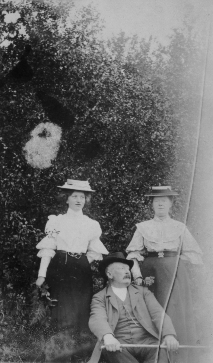 Tårnelv 1905. En mann sitter og to damer står på hver side av han. De står og sitter foran et stor tre eller busk.