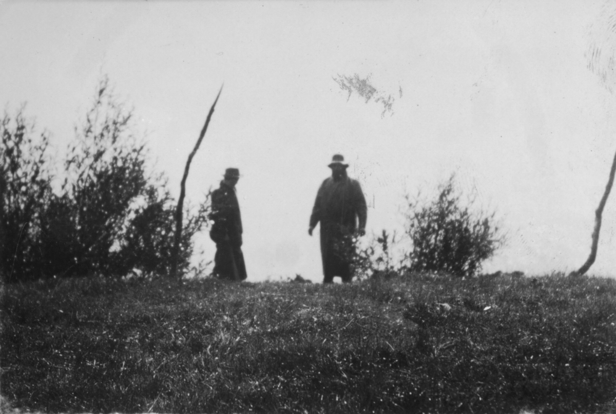 To menn står på bakken. Begge i hatt og frakk