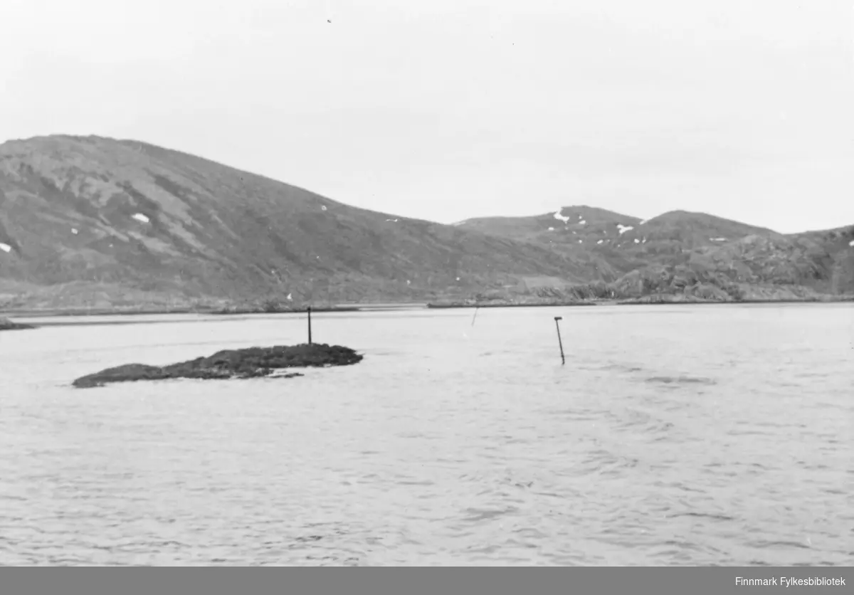 Naturbilde. Tatt utover fjorden i Tufjord. En åskam strekker seg utover fra venstre billedkant. Sjøen ligger rolig. Det er overskyet.Ute på fjorden kan vi se et skjær. Noen staker stikker opp av sjøen. Sikkert for å markere skjærene.
