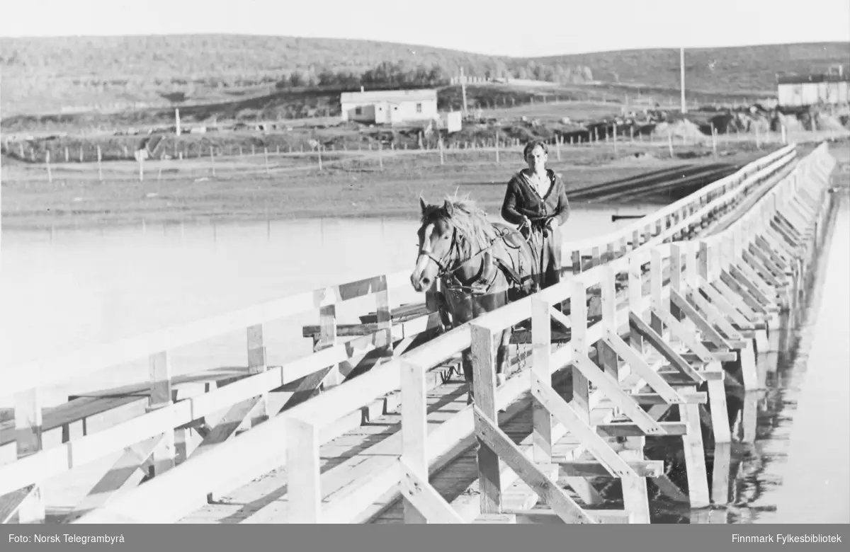 'Den provisoriske broen over Kautokeinoelven. Den er bred nok for ett hestekjøretøy. Høsten 1946.' En hest og vogn er på vei over elven. Det står en mann bak på vognen.