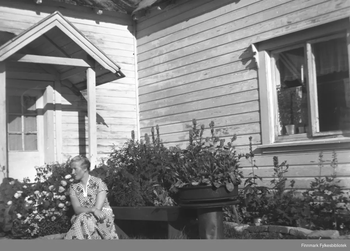 Herlaug Mikkola sittende i den lune kroken ved inngangspartiet på gården Mikkelsnes. Bak henne ser vi flotte bed med blomstrende hageblomster. I vinduet står det frodige potteplanter