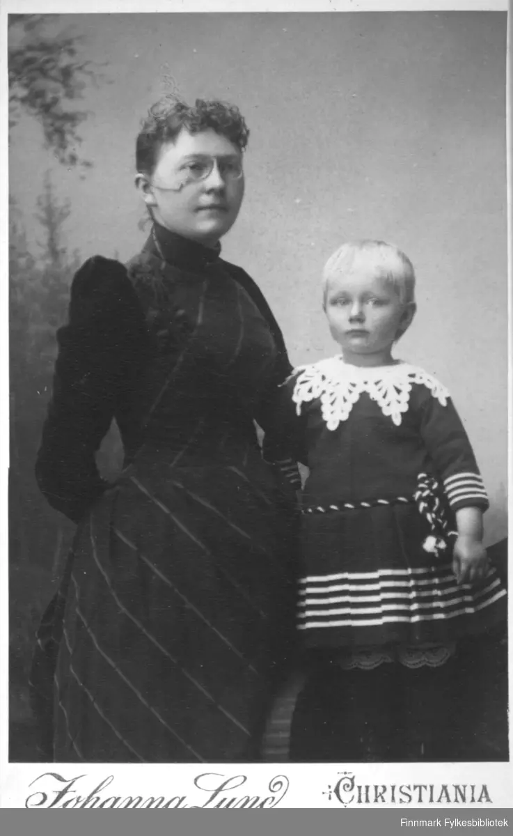 Portrett av en kvinne og et barn. Kvinnen har en mørk kjole på seg og lorgnetter på nesen. Barnet har en mørk drakt/kjole med striper rundt livet og håndleddene. En heklet krage ligger rundt halsen.