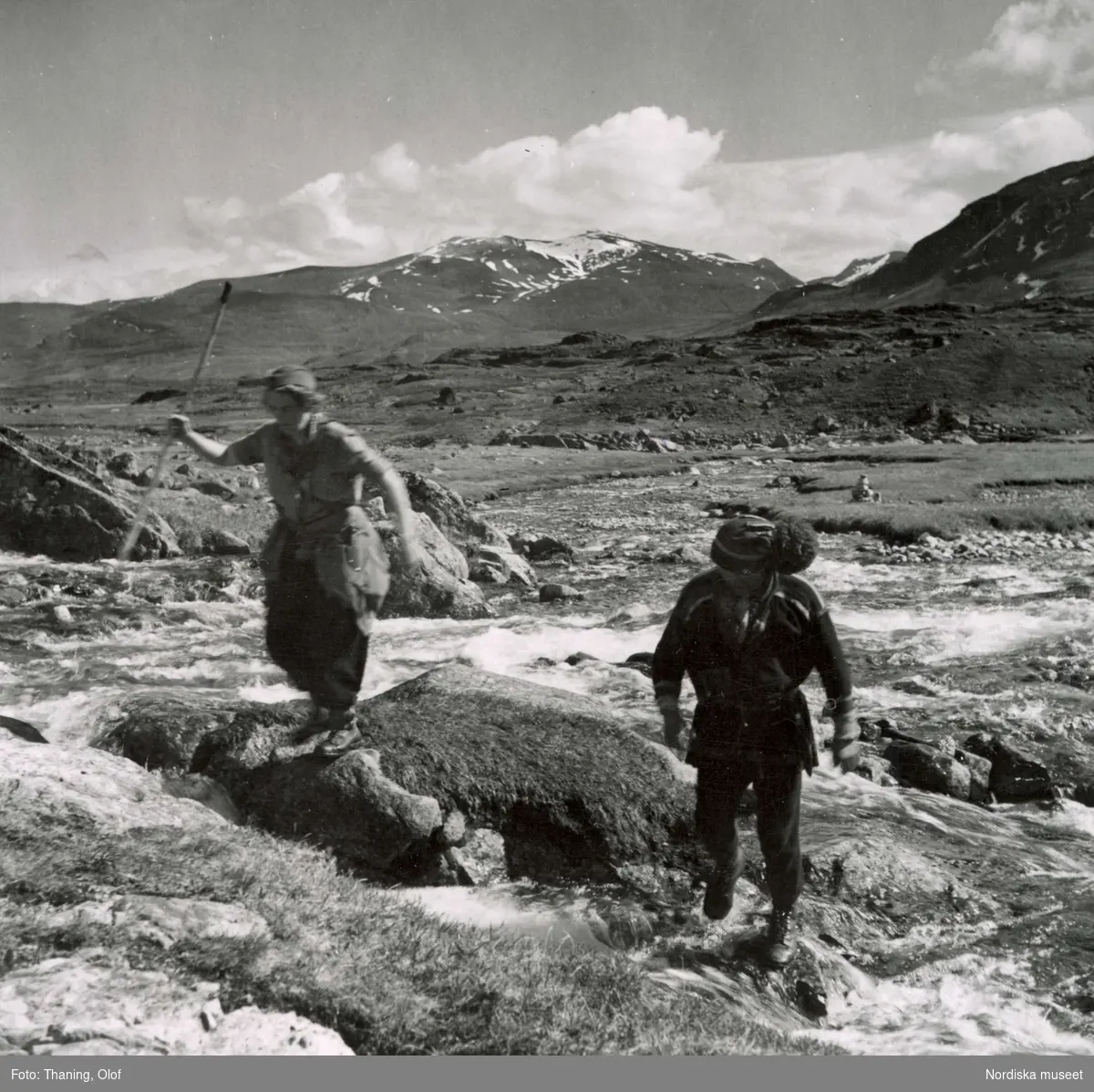 STF:s pressresa på Kungsleden. Kvinna med vandrarstav och samisk man tar sig över ett vattendrag.
