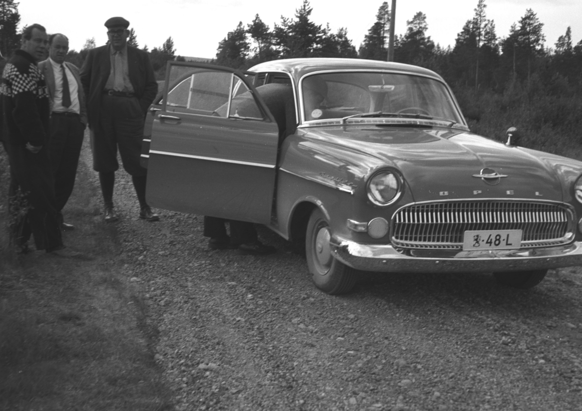 Tre personer står ved siden av en bil, Opel Kaptein årsmodell 1956-57. Det tilsier at bildet tidligst kan være tatt høsten 1955. Registrert på uvanlig finsk skilt, med den finske riksløven først og bokstaven bakerst. Tyder på at den er statlig, med spesiell status, kanksje "Riksdagsbesøk".