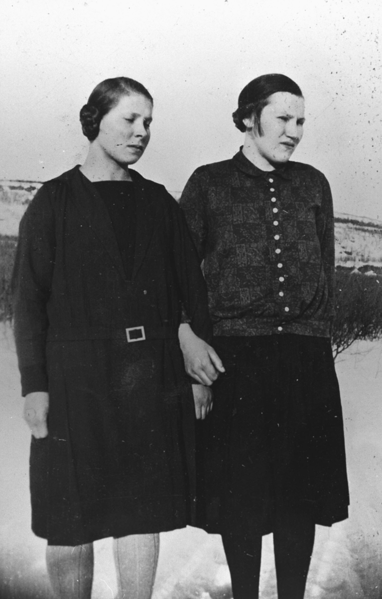 To unge kvinner, (fra venstre) Elna Mietinen (født Malmstrøm) og Emma Aleksandersen, fotografert ute på en vårdag rundt 1930. Kvinnene holder hverandre i hånden, Elna har lukket igjen øynene sine fordi det er skarp lys ute. Det er fortsatt snø på bakken. Bildet er tatt fra nært avstand.