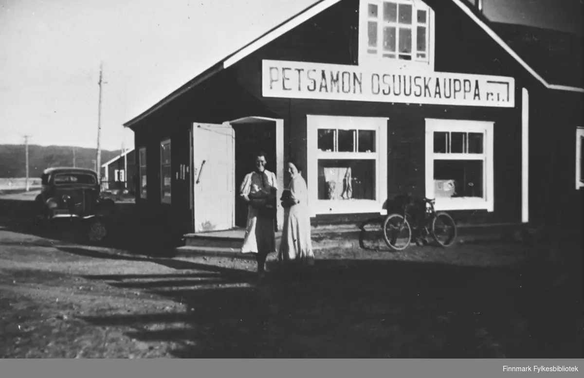 Et par fotografert utenfor butikken / kolonialhandelen  i Petsamo / Petsjenga. Biler og sykler står parkert utenfor
