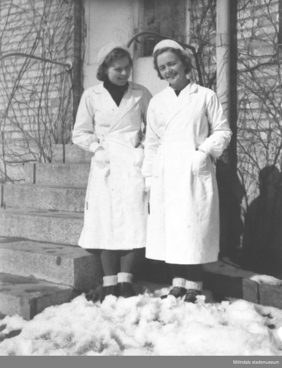 En okänd kvinna och Rosa Pettersson (född Krantz) står klädda i sina vita arbetsrockar, okänt årtal. Kvinnorna står i snön utanför ett charkuteri i Solängen.