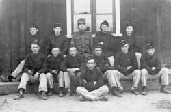 Tolv unge menn samlet ved husveggen, soldater framfor ei kas