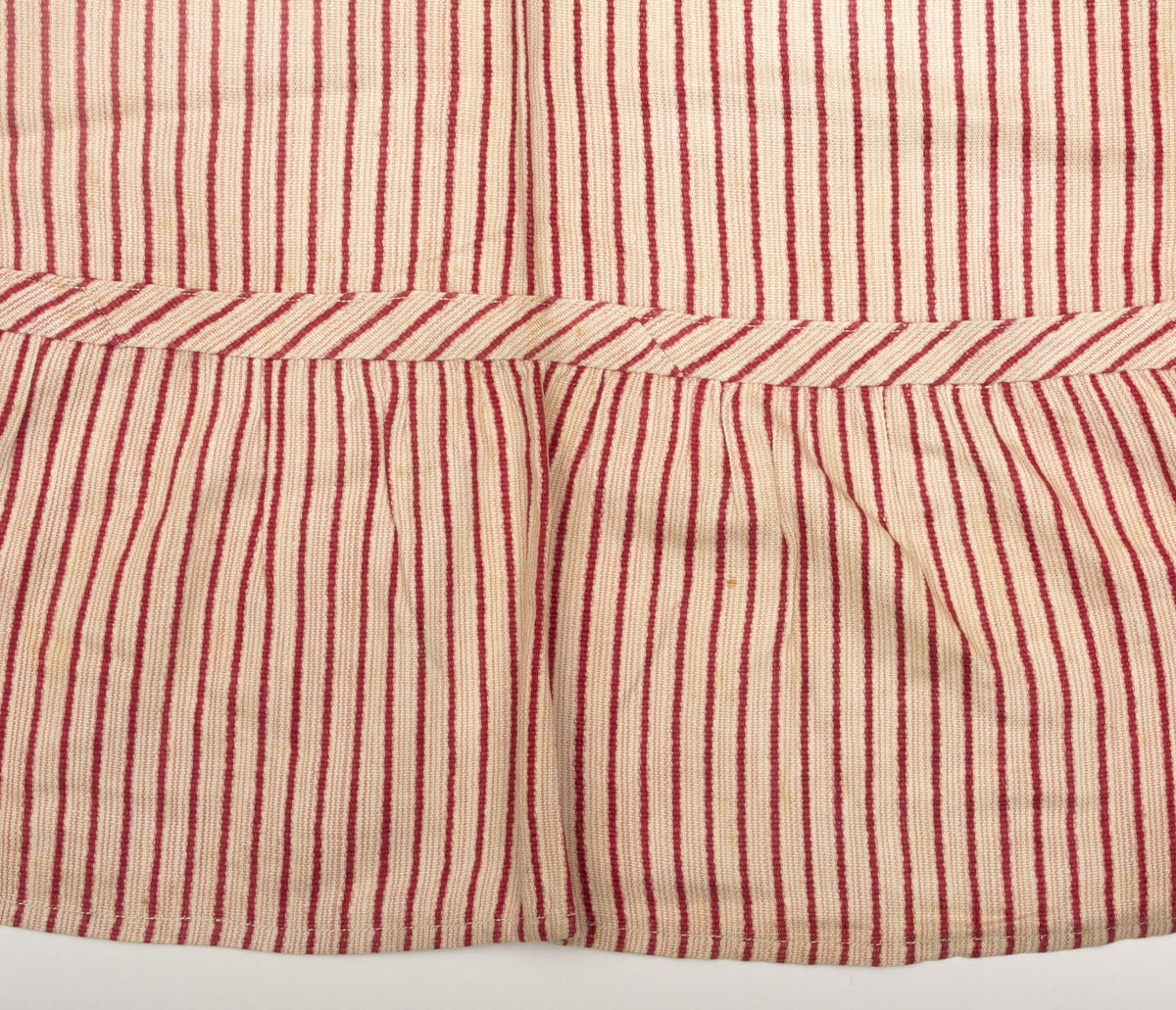 Långrandigt bomullsförkläde med smala rosa ränder på vit botten, varje cm en något bredare röd ripsrand. Nertill en 16 cm bred volang. Linning och knytband av tyget.
