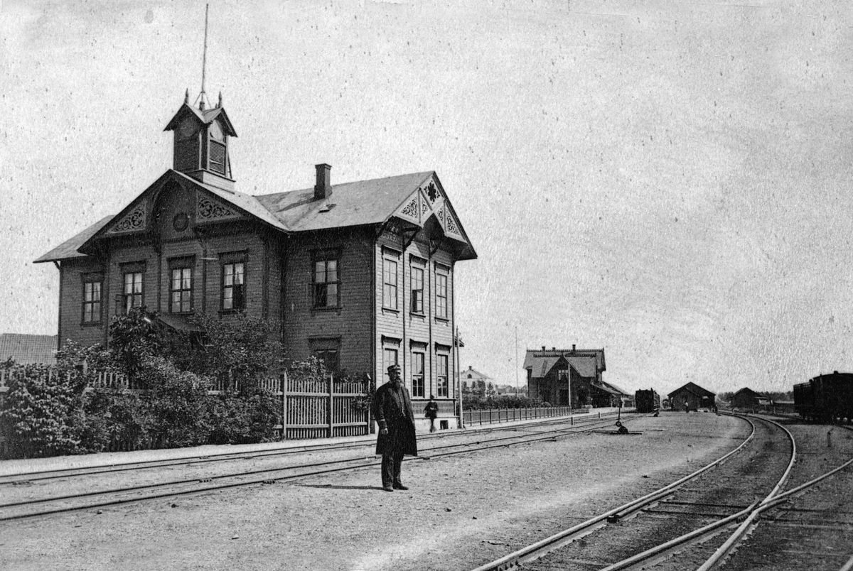 Hamar jernbanestasjon, Hamars første stasjonsbygning fra 1862, åpnet 23. juni, Grundsetbanen Hamar- Elverum, i bakgrunnen sees Hamars andre stasjonsbygning som ble tatt i bruk i 1880 for Hamar-Eidsvoldbanen, i forgrunnen ingeniør/stasjonsmester Christian Fredrik Lund (1838 -1886), (gift med Anne Christine f. Amundsen)