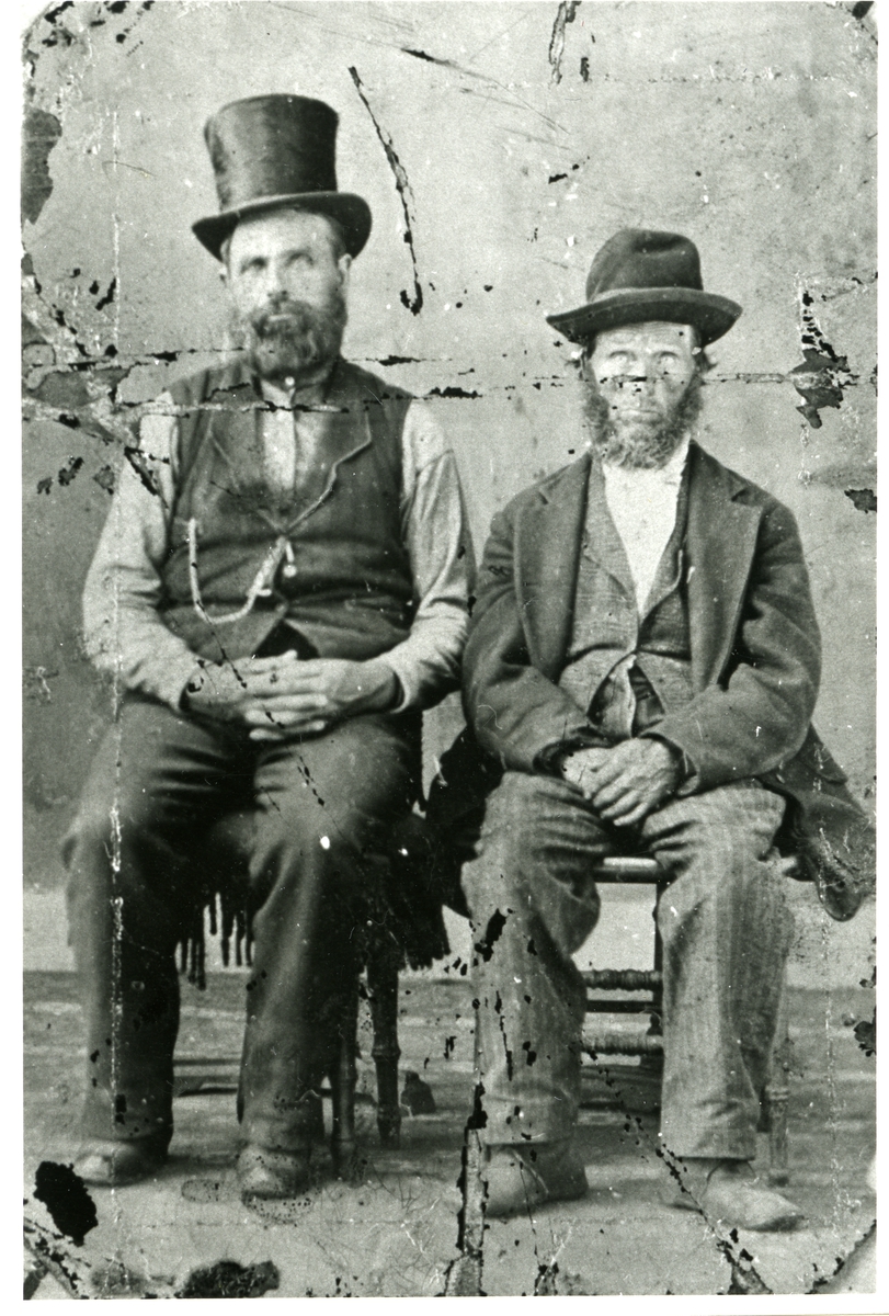 Portrett av to menn sittende på hver sin stol. Mennnene er begge iført dress og hatt.