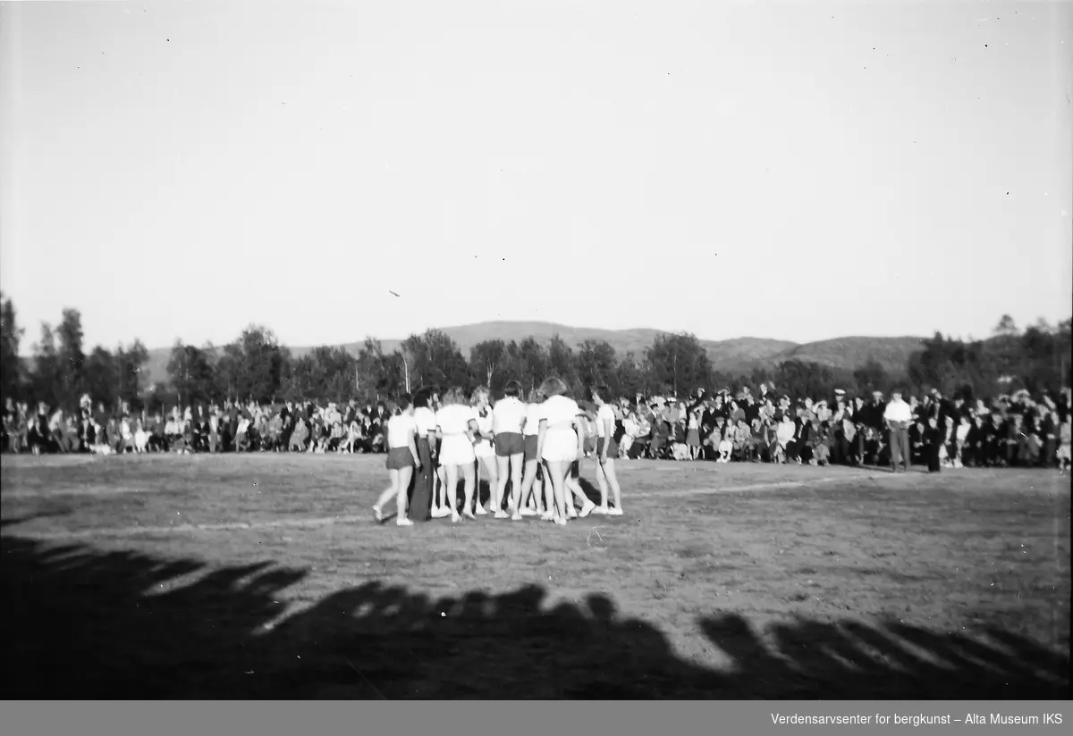 Et kvinnelag samles på banen før en fotballkamp. En stor folkemengde står ved sidelinjen og ser på.
