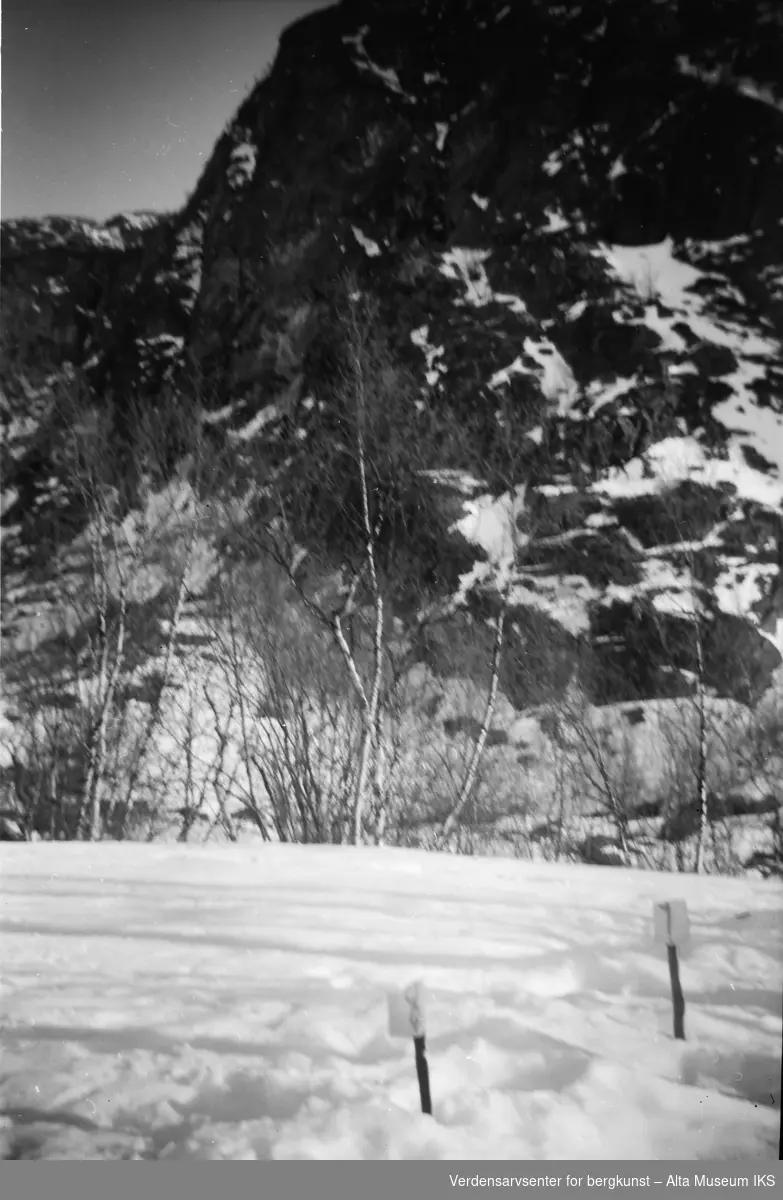I snøen står det pinner som markerer lengde, antakeligvis brukt til skihopping. I bakgrunnen tårner Raipasfjellet.