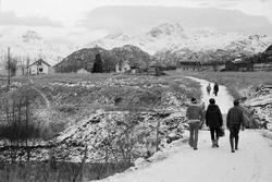 Fem personer går oppover en snødekket vei. Gårdsbebyggelse o